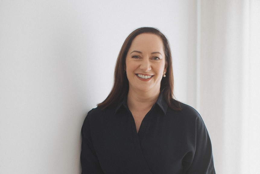 Dr Anita Collins, a white woman in the 50s with brown hair, is wearing a black dress and leaning against a white wall, smiling.