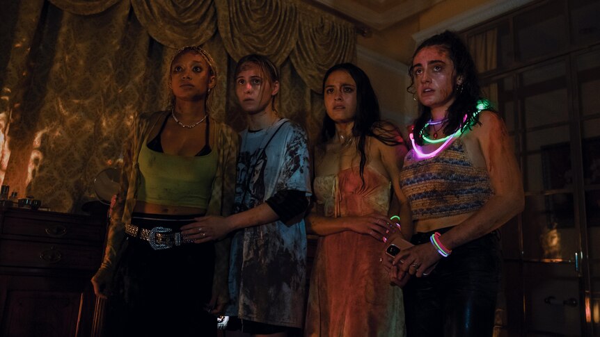 Four women splattered with blood, stand in a room in the semi-darkness looking concerned.