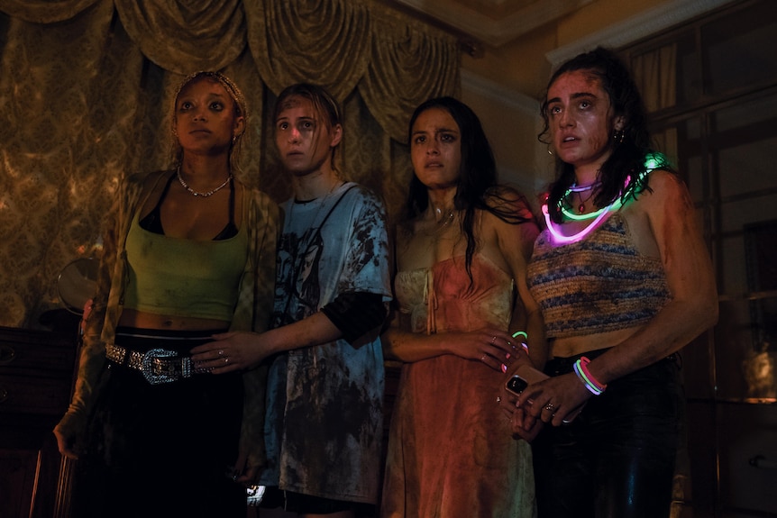 Four women splattered with blood, stand in a room in the semi-darkness looking concerned.