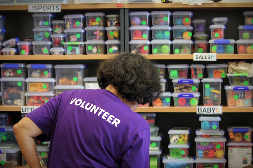 A woman in a purple volunteer shirt stares at a wall of toys in small plastic boxes 
