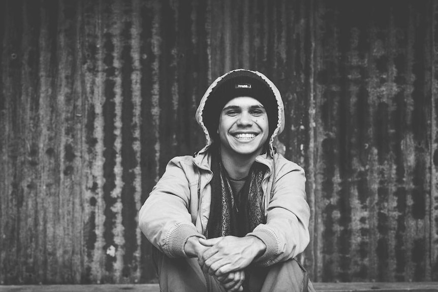 Portrait of a young Yaegl man sitting on a curb with a metal corrugated wall behind him.