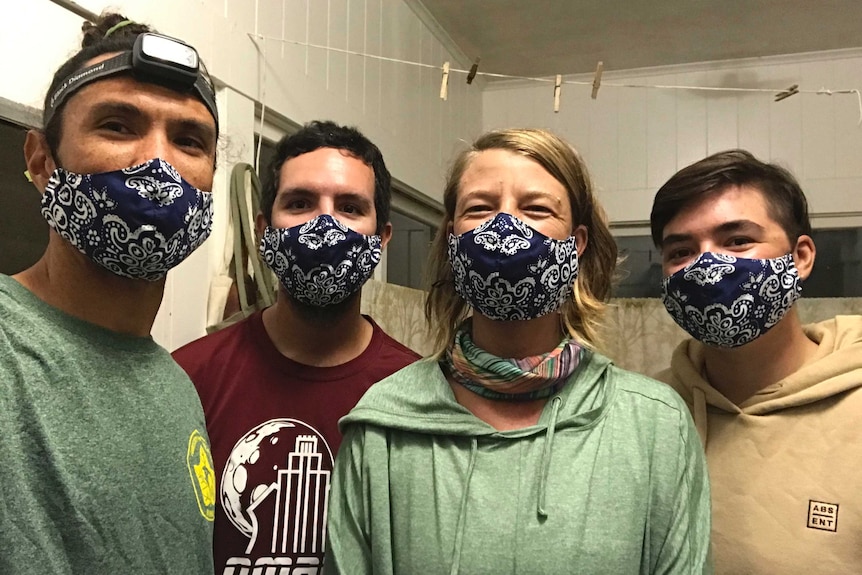Four people are seen in a room at what appears to be night wearing matching blue patterned face masks and smiling.
