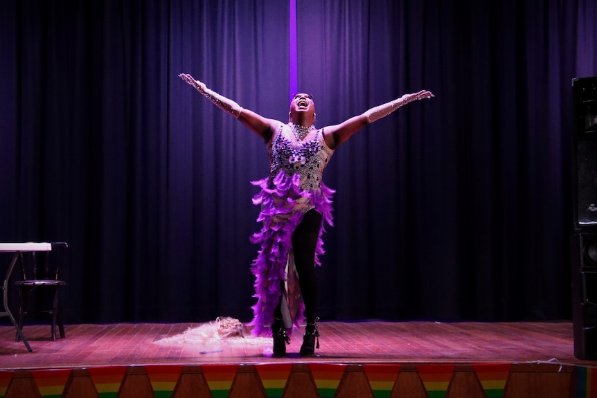 A man in drag has his arms out, singing on stage. 