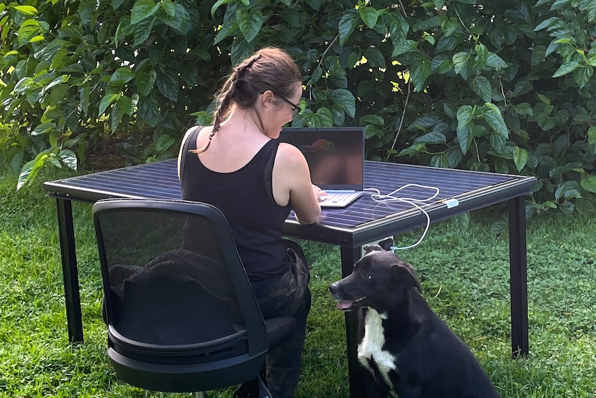 A woman sits at a table made out of a solar panel with a dog next to her in a garden.