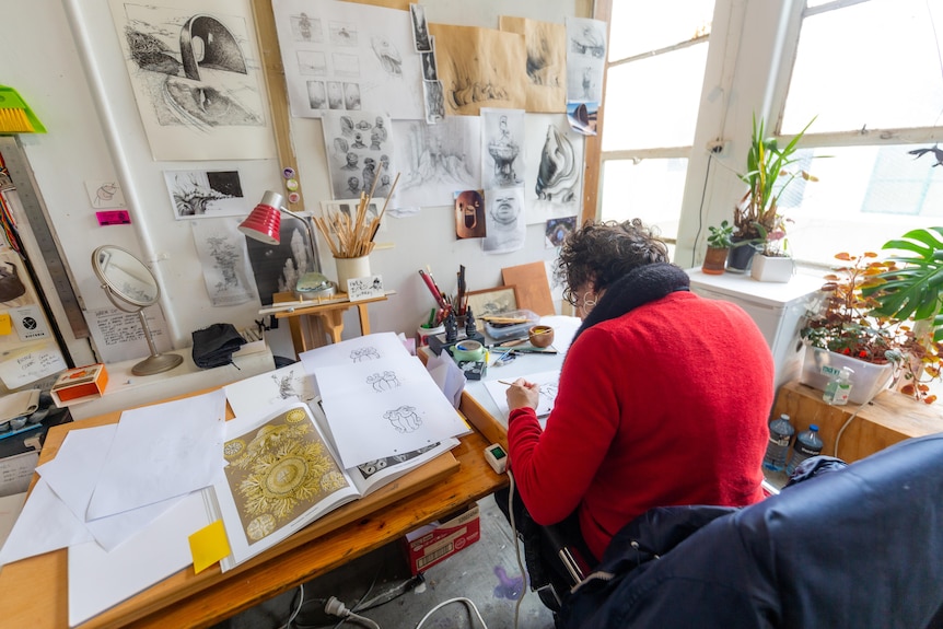A woman sitting at a desk surrounded by drawings with her back to the camera.