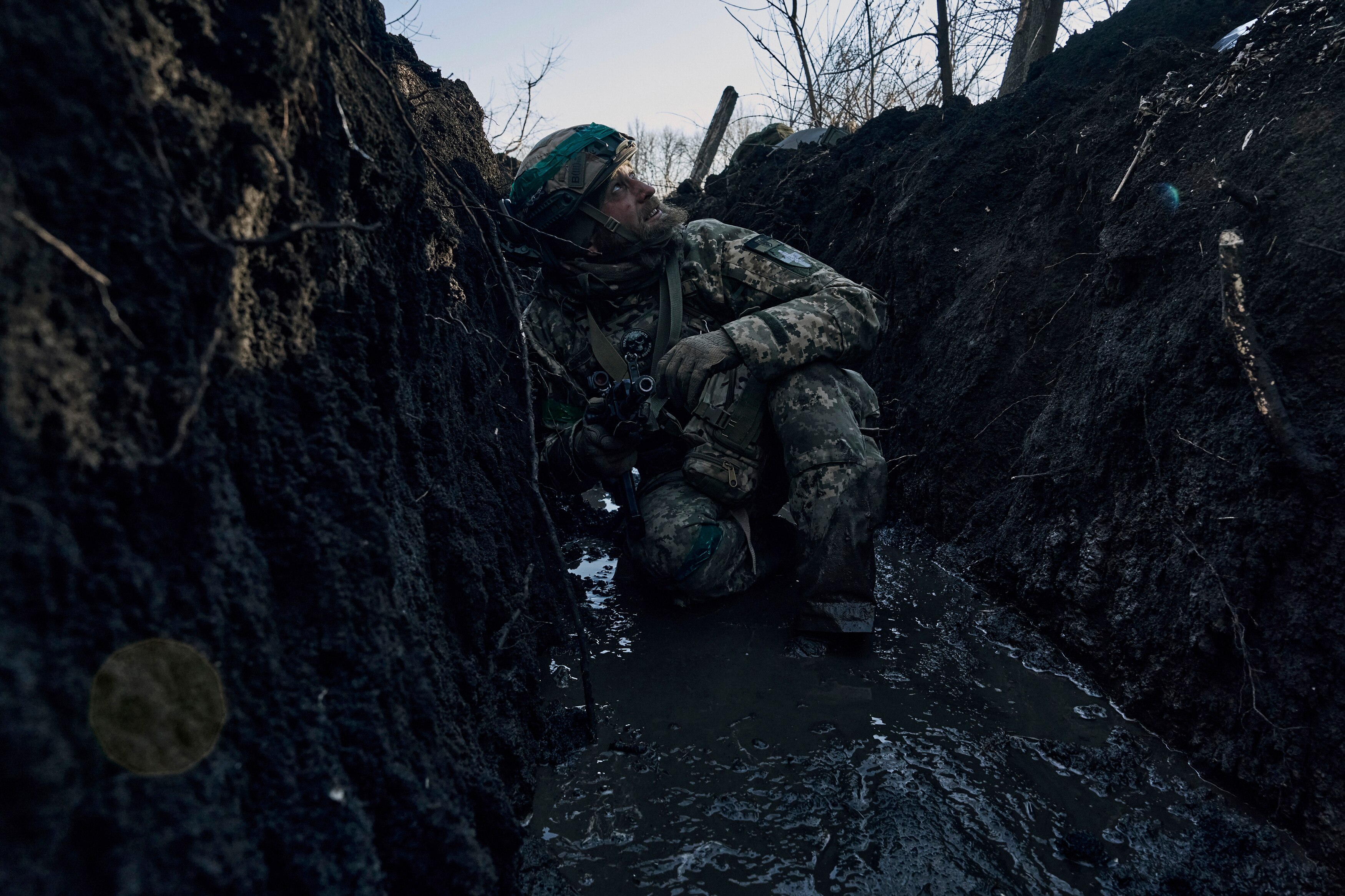 A bearded man in a soldier's combat uniform squats in a trench filled with mud as he looks up, concerned, at the sky above.