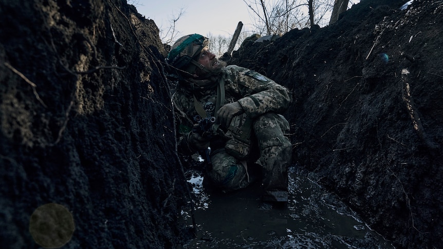 A bearded man in a soldier's combat uniform squats in a trench filled with mud as he looks up, concerned, at the sky above.
