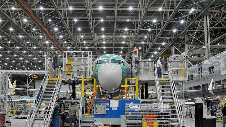 In a large air hangar, a green Boeing plane is surrounded by scaffolding about two storeys high with plenty of machines beside.