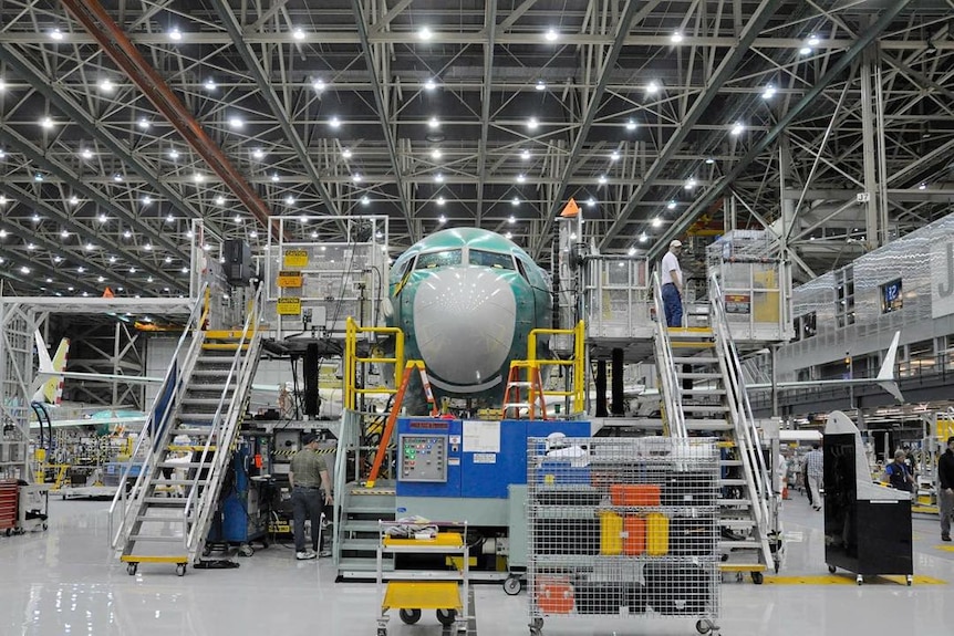 In a large air hangar, a green Boeing plane is surrounded by scaffolding about two storeys high with plenty of machines beside.