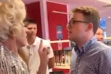 A young man stands shouting at a drag queen, covered in pearls, in a library. The queen is holding her finger to say 'stop'.