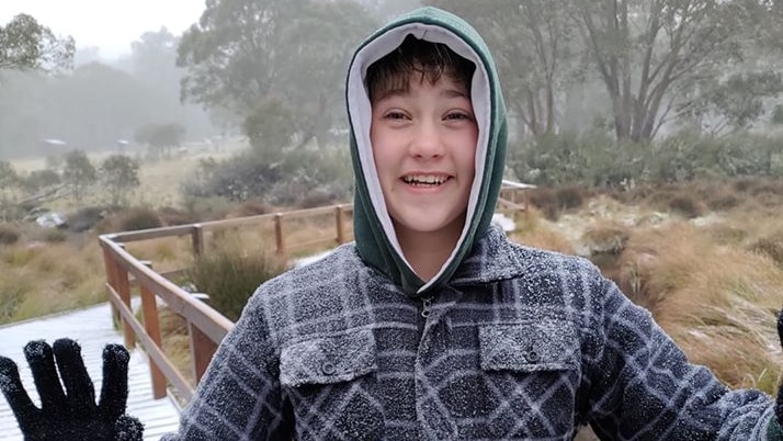 A boy dressed in winter gear grins, with his hands outstretched amid snow falling on a bush track.