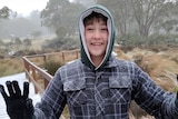 A boy dressed in winter gear grins, with his hands outstretched amid snow falling on a bush track.