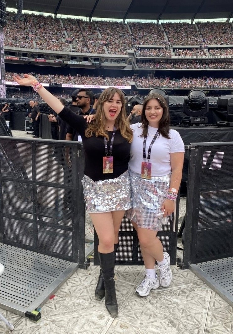 Dos mujeres de veintitantos años con faldas plateadas brillantes sonríen dentro de un estadio.