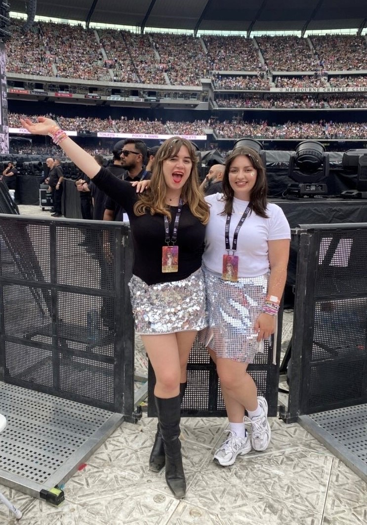 Two women in their twenties in sparkly silver skirts smile inside a stadium.