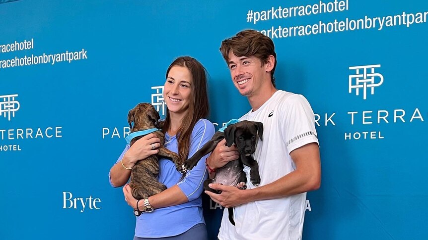 Two tennis players - one female, one male - dressed in casual clothing stand in front of signs for an event while holding dogs.