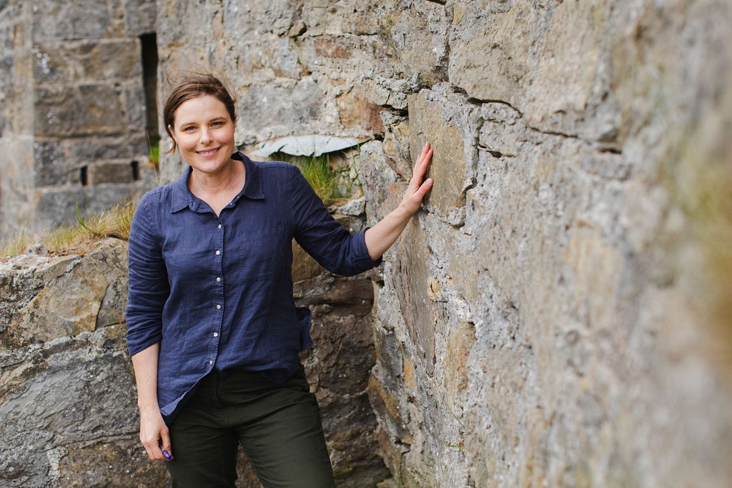 A smiling woman stands next to an old wall, resting her hand against it