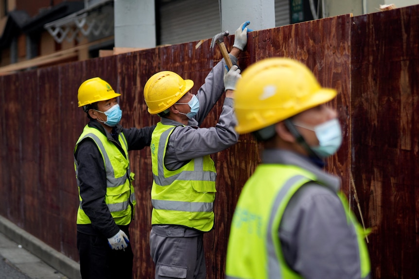 Three workers in hard hats and high vis vests use hammers to erect sheets of timber barriers. 