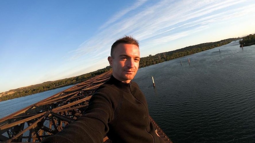 A man in a black jacket takes a selfie at the top of a high rise building. 