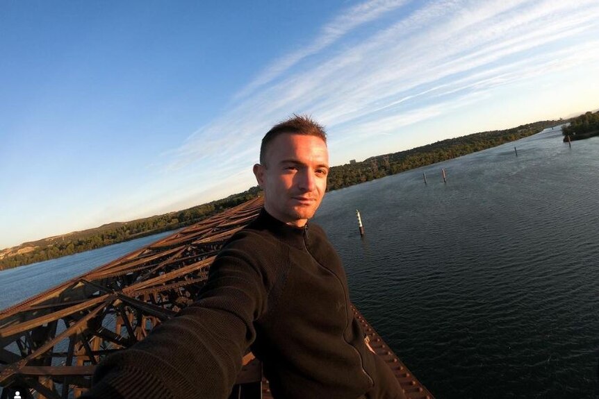 A man in a black jacket takes a selfie at the top of a high rise building. 