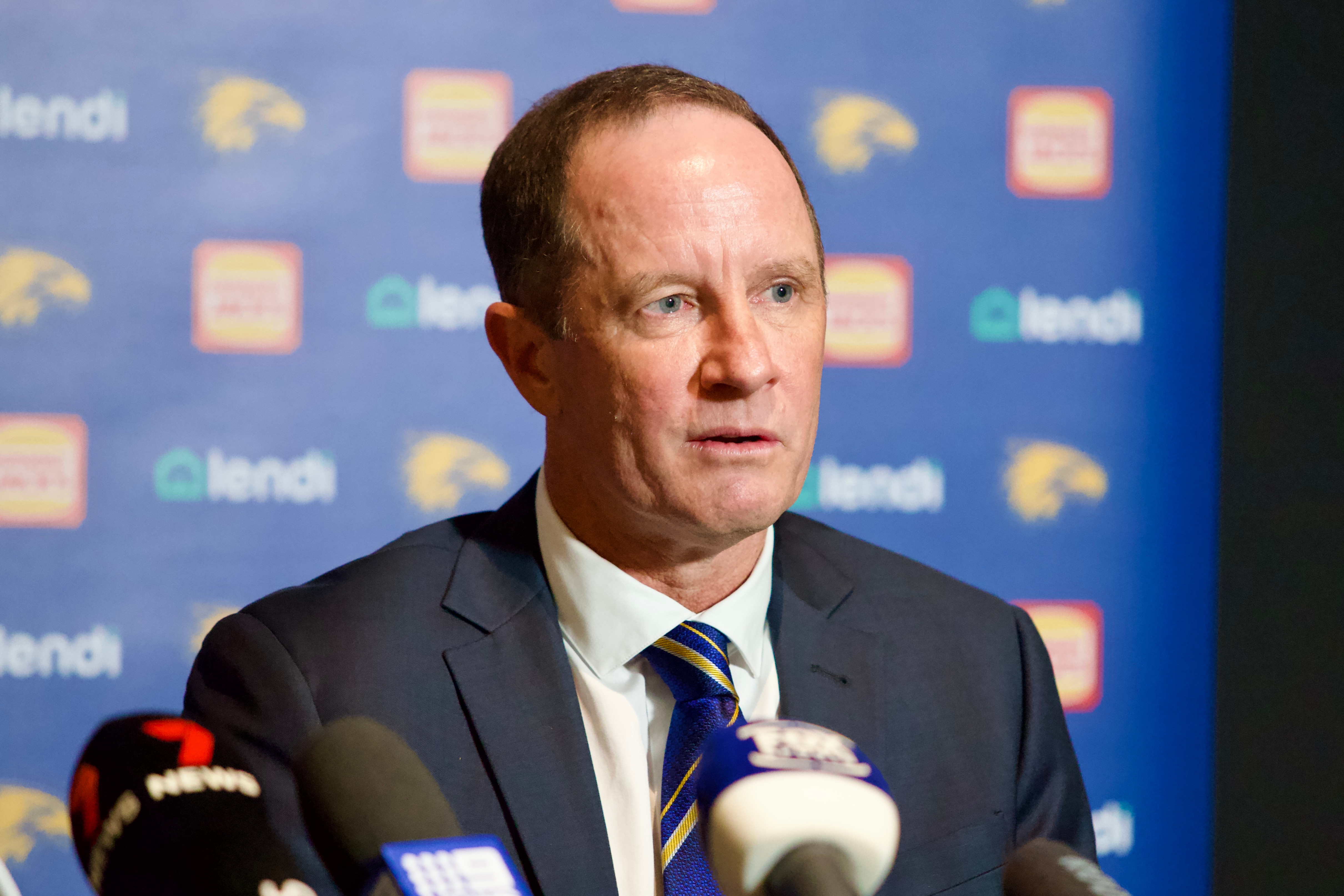 A man in a suit and tie is sitting and speaking to the media at a press conference with a branded banner behind him.