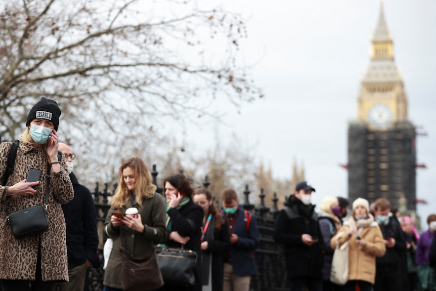 A group of people, some wearing face masks and others without, line up outside. Elizabeth Tower, AKA Big Ben, is visible behind