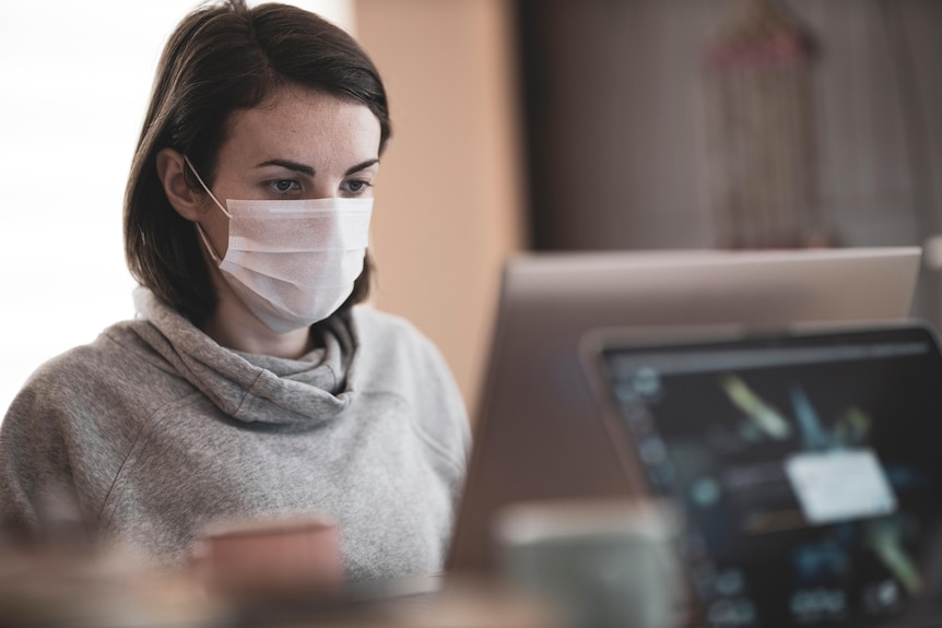 A woman wears a white mask while working at a computer.