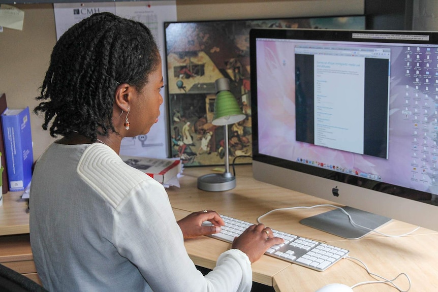 A woman of African descent sits at a desk in front of a computer screen
