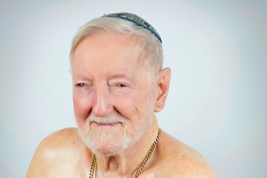 An older man wearing a blue kippah (jewish hat), and two gold chains, one with the star of David. Shirtless, white beard