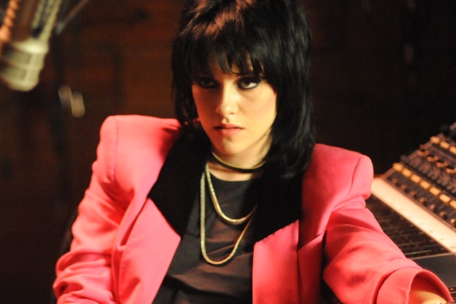 A young woman with 80s style punk-goth make-up and hairdo and bright blazer sits looking unimpressed in recording studio.