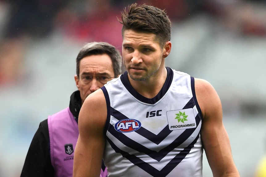 A Fremantle Dockers AFL player walks on the MCG playing surface with a trainer walking behind him.