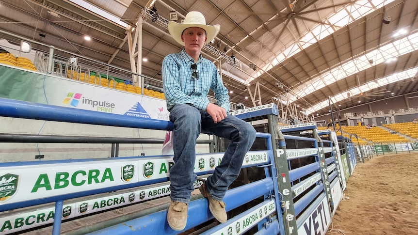 A boy in a white hat and blue shirts sits atop a rodeo chute.
