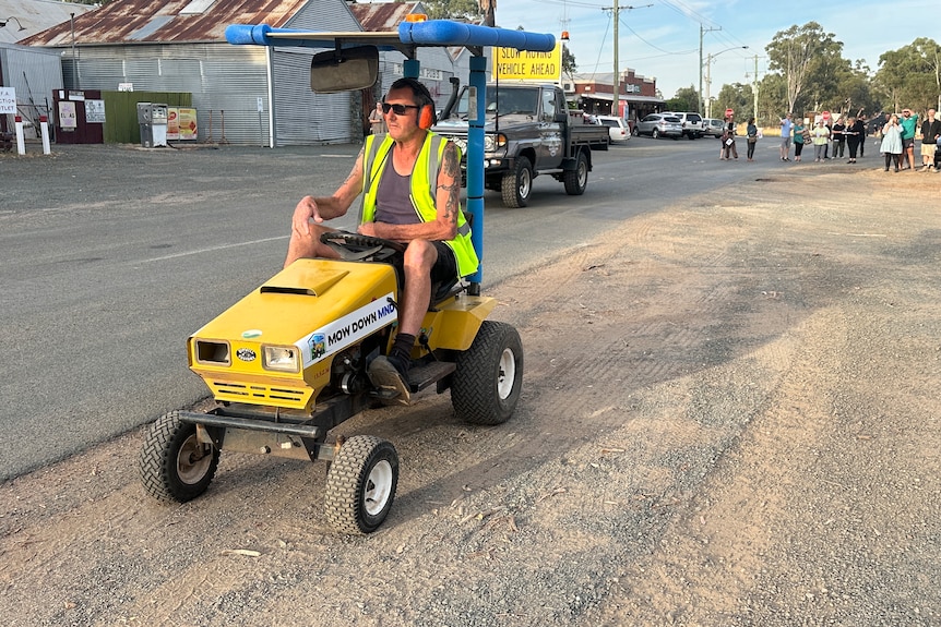 A man in high-vis steers a ride-on mower down a dusty street.