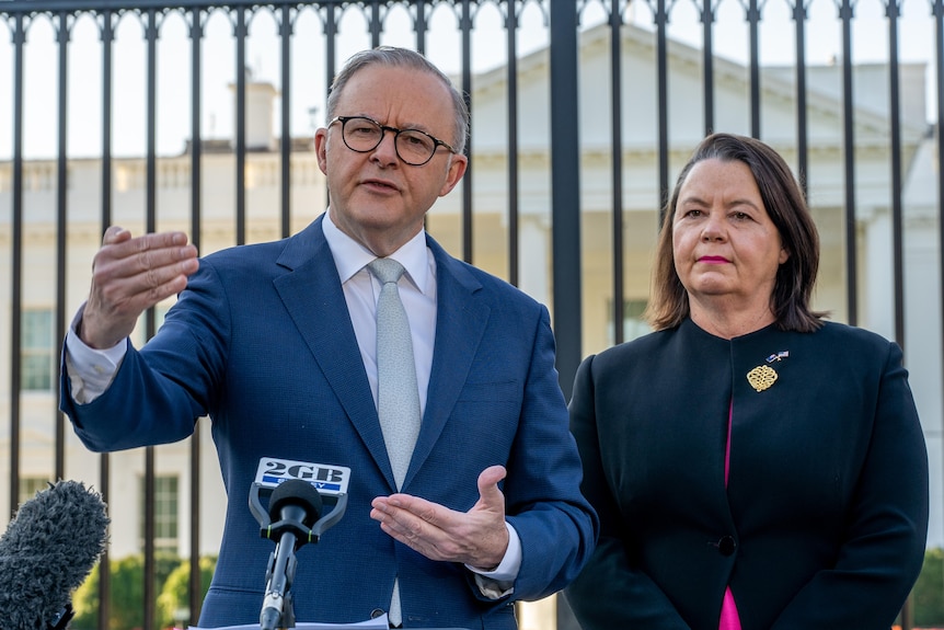 Anthony Albanese speaks at a podium with Madeleine King behind him by a fence in front of the White House.