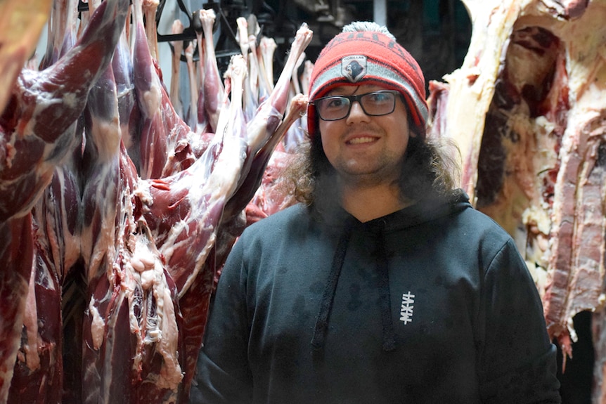 A man in a hat smiles next to wallaby carcasses hanging in meat factories. 