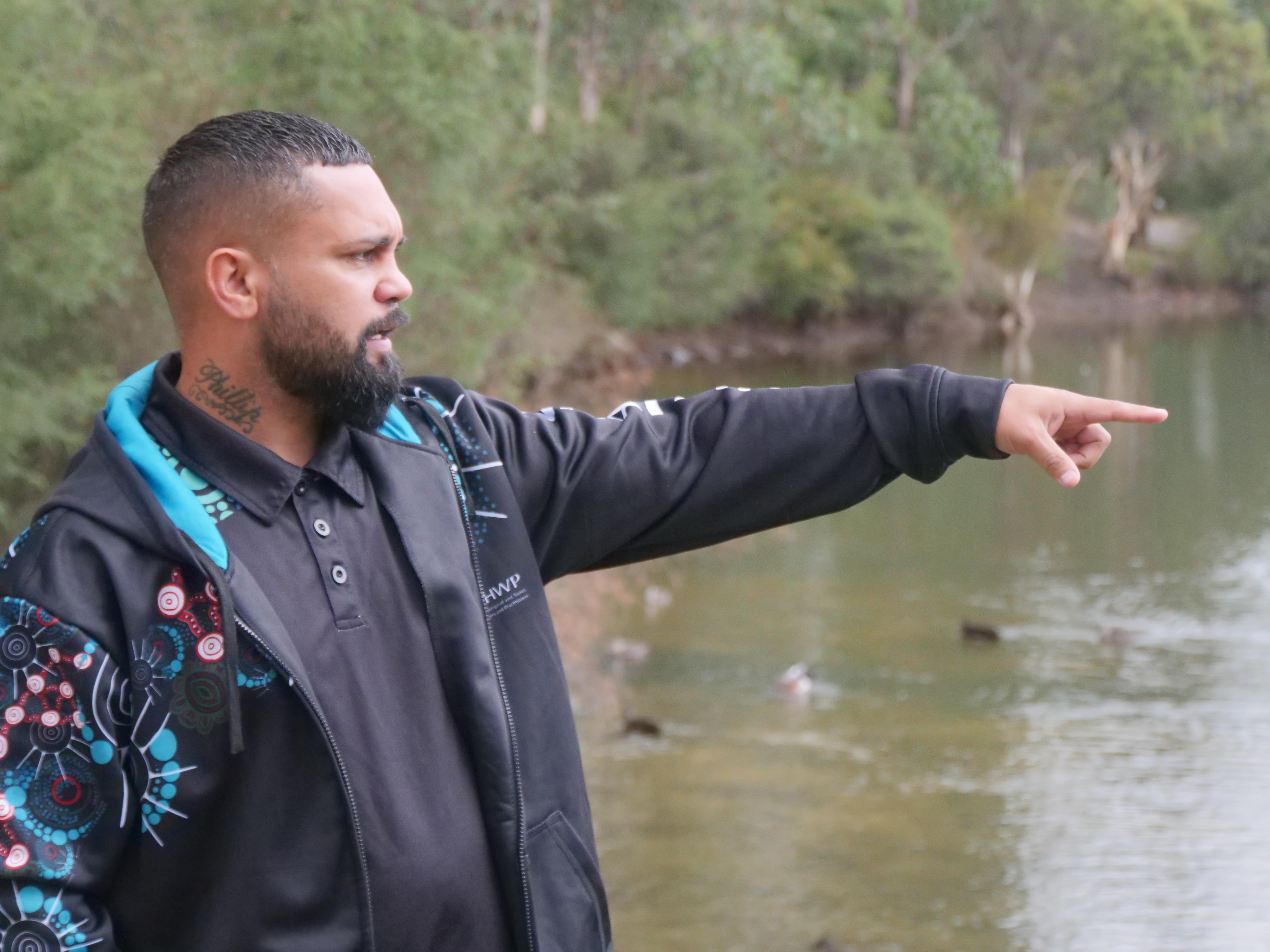 An Indigenous man in a dark jacket points to something unseen as he stands next to a waterway.