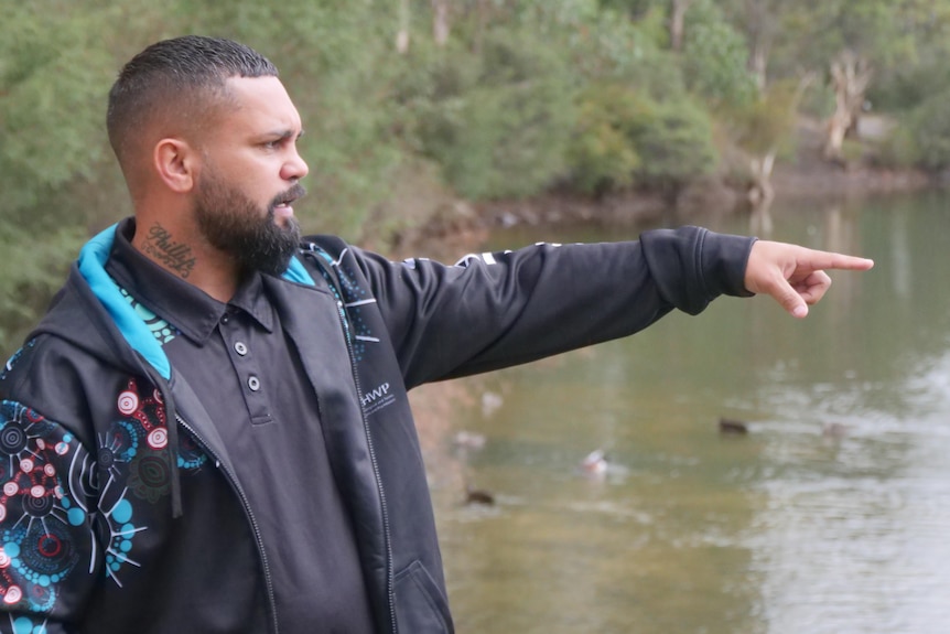 An Indigenous man in a dark jacket points to something unseen as he stands next to a waterway.