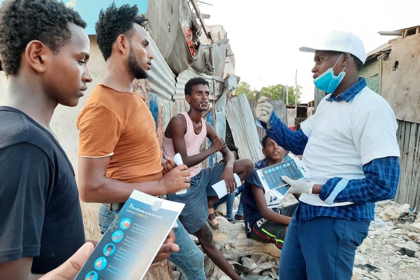 An IOM representative distributes pamphlets to four men outside some rundown housing.