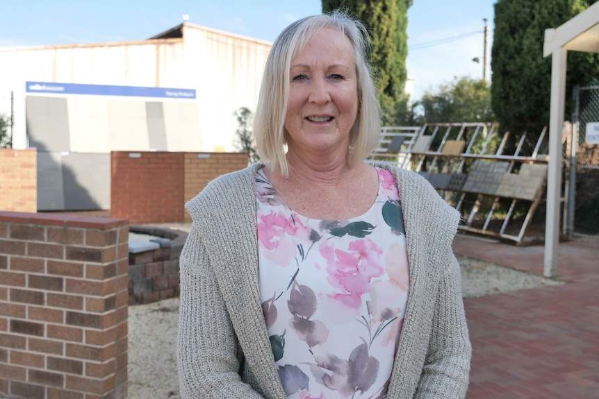 Woman with blonde hair, beige cardigan and pink floral top in front of a display yard of pavers and tiles.