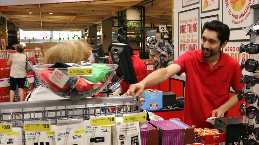 A man in a red shirt smiles as he serves a woman across a shop counter.