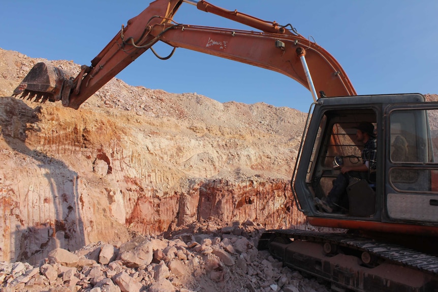 A man in a blue checked shirt controls an excavator, which is removing hard sand-coloured stone from a wall deep in the ground.