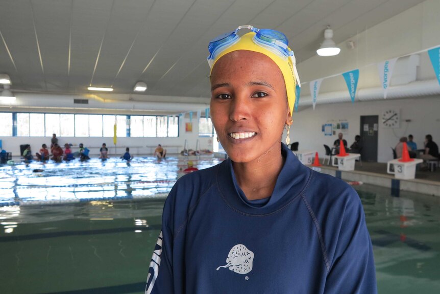 A young African woman smiles in tracksuit and swimming cap in an indoor complex with pool behind her