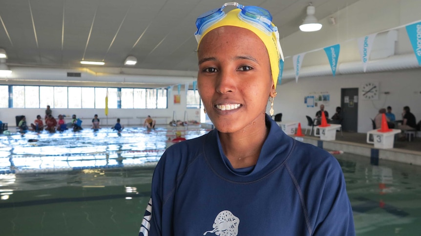 A young African woman smiles in tracksuit and swimming cap in an indoor complex with pool behind her