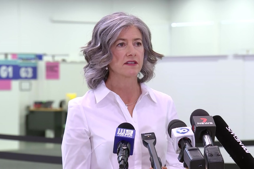 A grey-haired woman in a white shirt addresses the media. She is standing behind five microphones all pointed in her direction