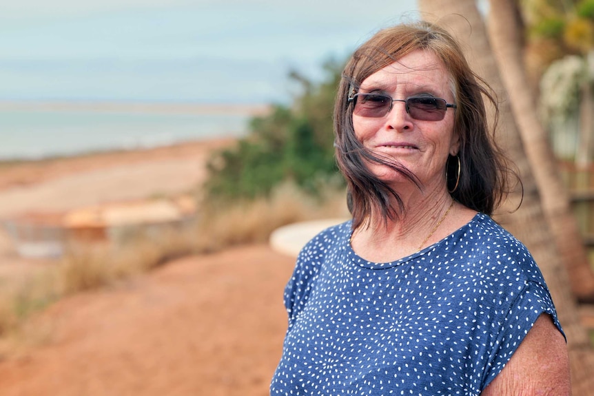 A woman wearing a blue shirt stands near the edge of the ocean and looks toward the camera.