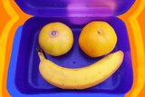 Two mandarins and a banana form a smiley face inside a blue plastic lunch box, to depict healthy lunchbox swaps for kids.