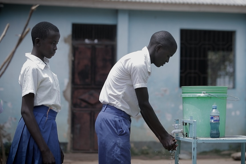 A school student washing his hands under a bucket with a tap, with another waiting their turn behind him.
