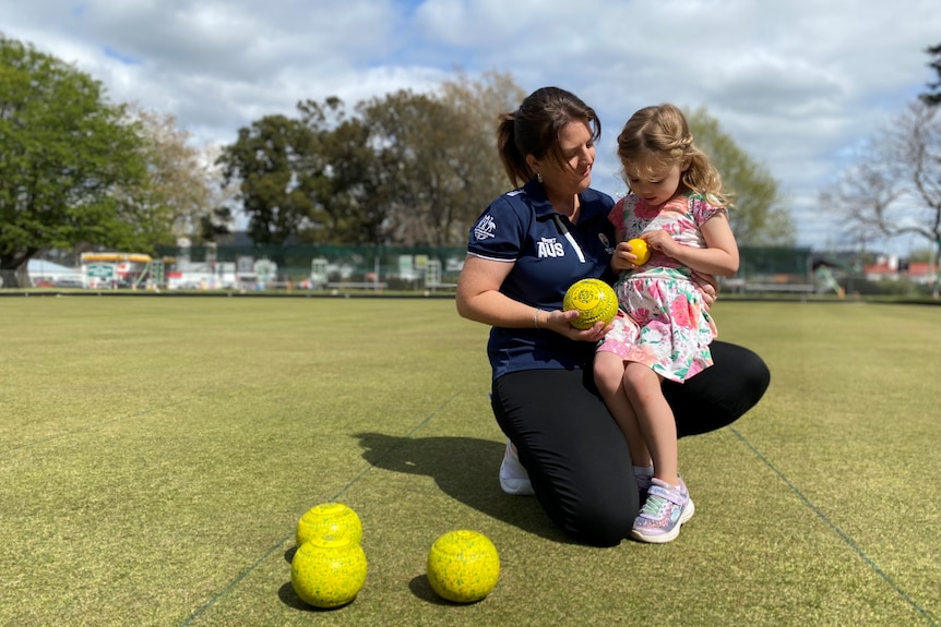 A little girls sits on a woman's knee as the pair look at lawn bowls, smiling.