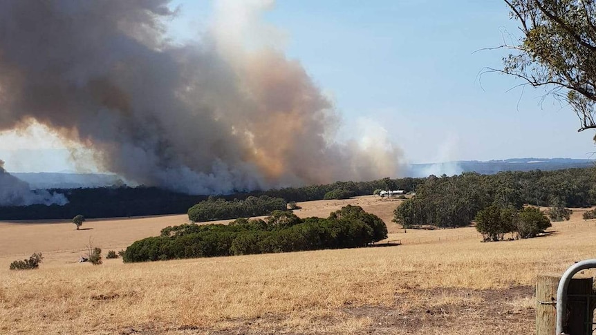 Orange-tinged smoke rises above dry paddocks of grass and bushland.