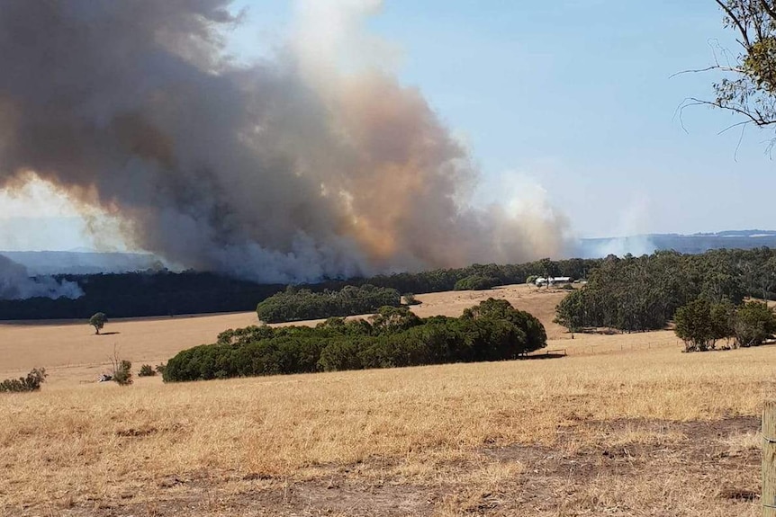 Orange-tinged smoke rises above dry paddocks of grass and bushland.