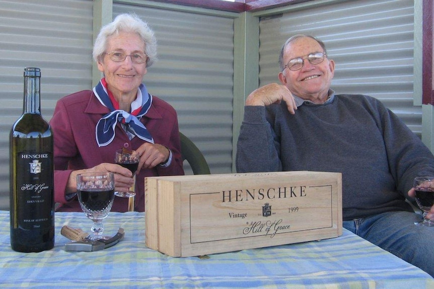 An elderly couple sit at a table, each with a glass of red wine in hand.